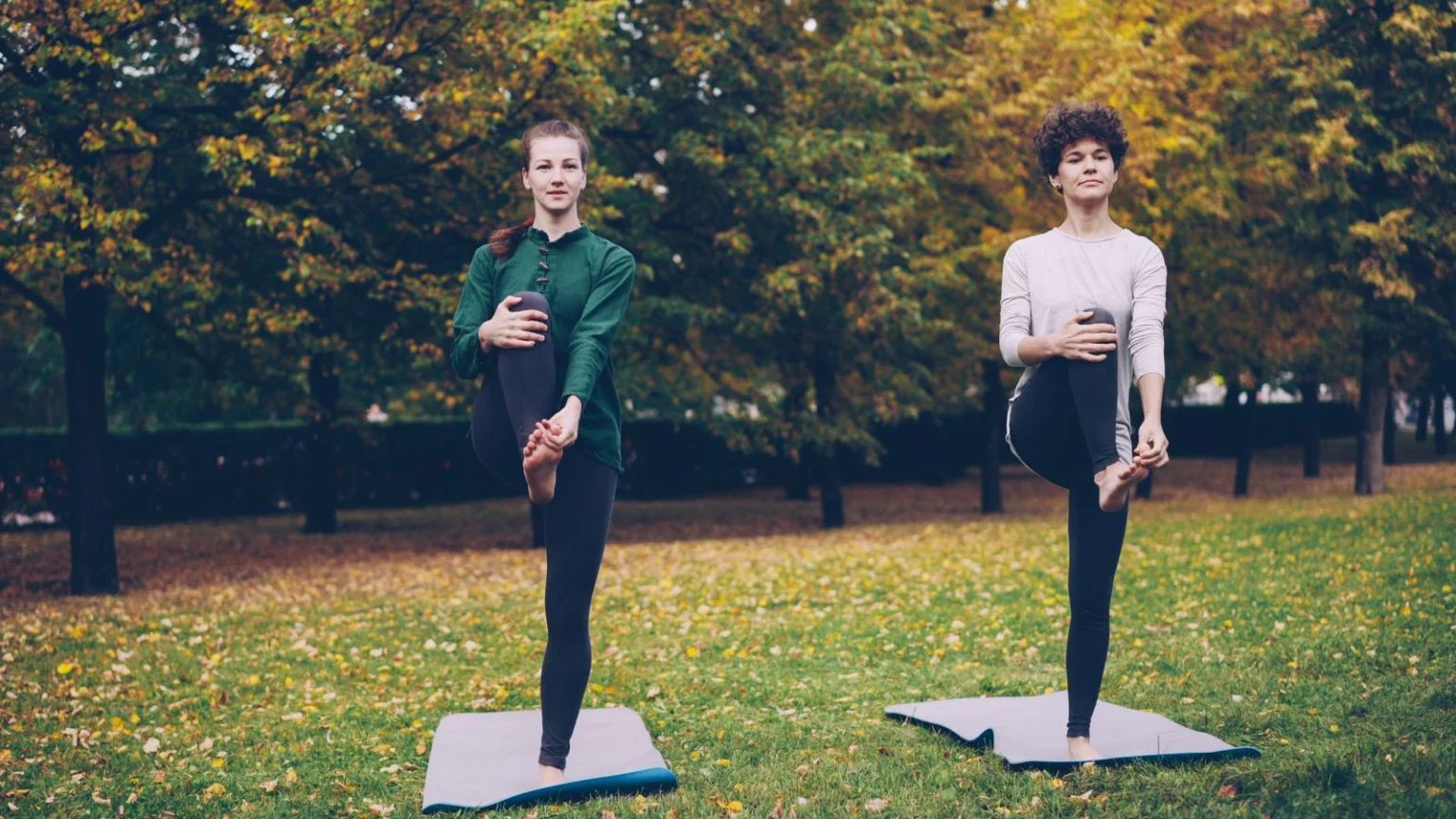 Two women doing a balance pose on yoga mats in a park